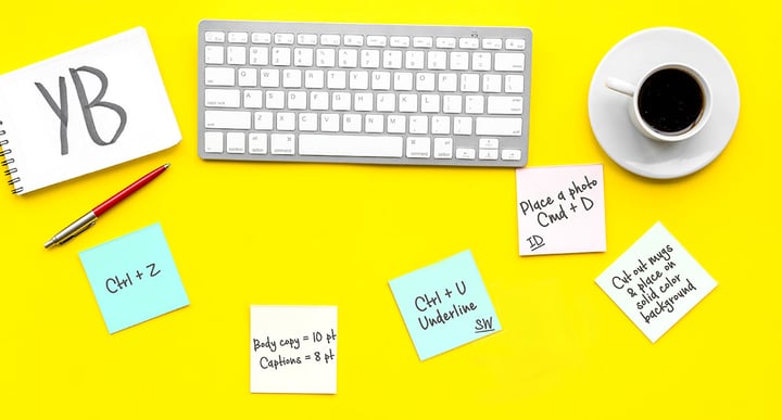 Yellow desk top including a cup filled with coffee, sticky notes, ink pen and wireless keyboard.